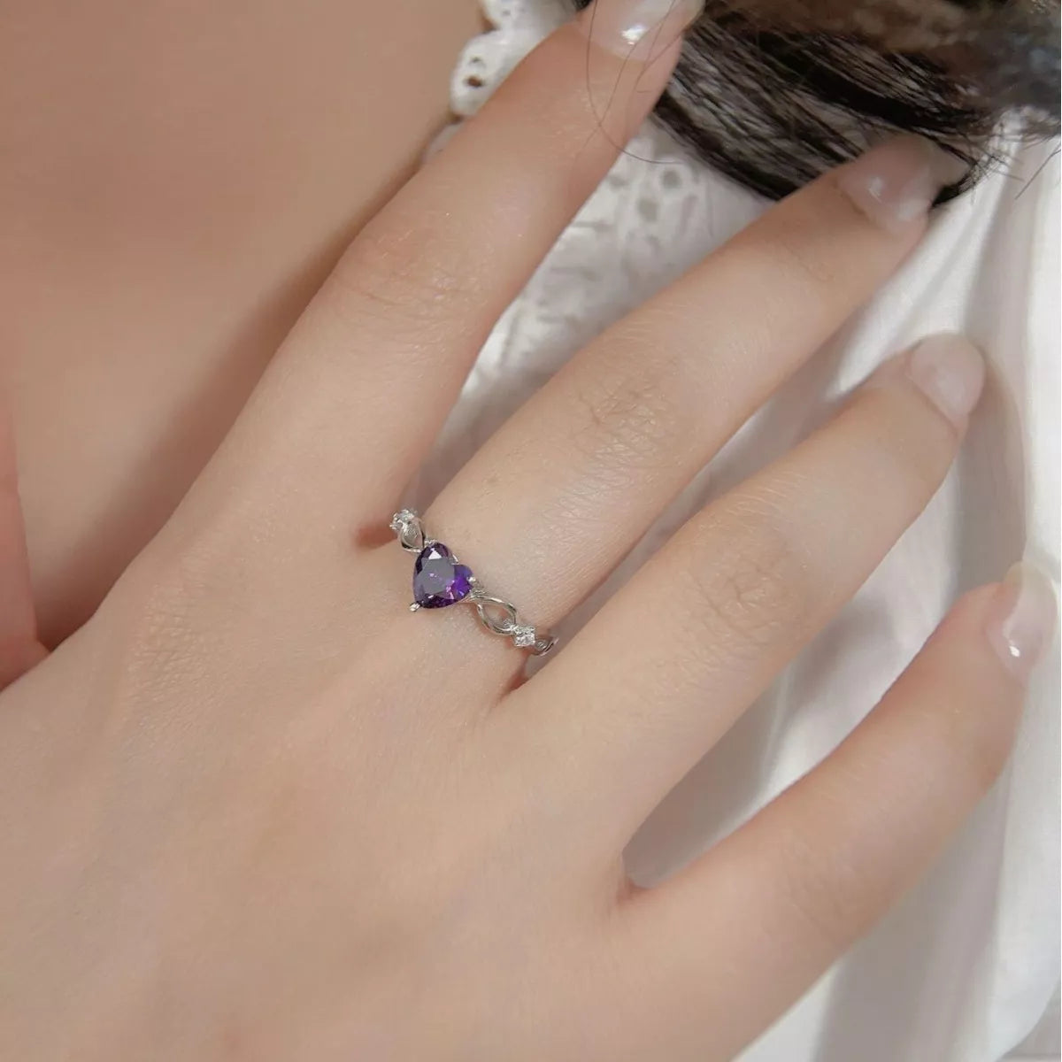 Silver ring with heart-shaped purple amethyst gemstone shown on a woman’s hand wearing a white blouse